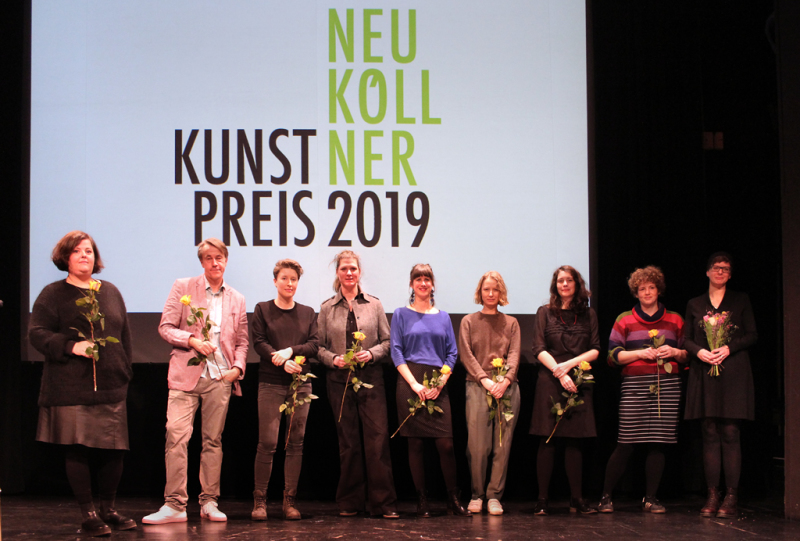 Neun Menschen stehen mit Blumen in der Hand auf einer Bühne vor einer Projektion der Schrift Neuköllner Kunstpreis 2019