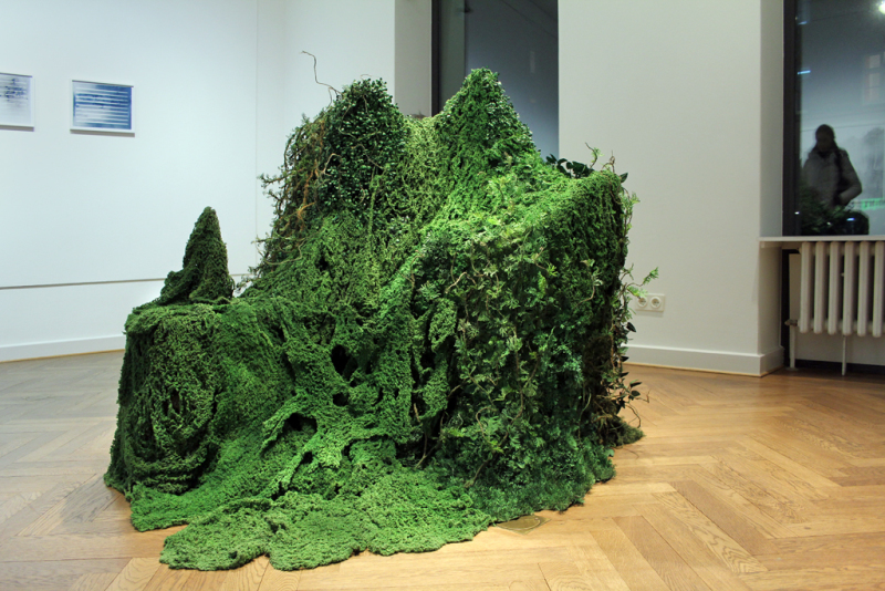 Eine Skulptur bestehend aus von künstlichen Pflanzen überwachsenen Gartenmöbeln, Flaschen und Gläsern.