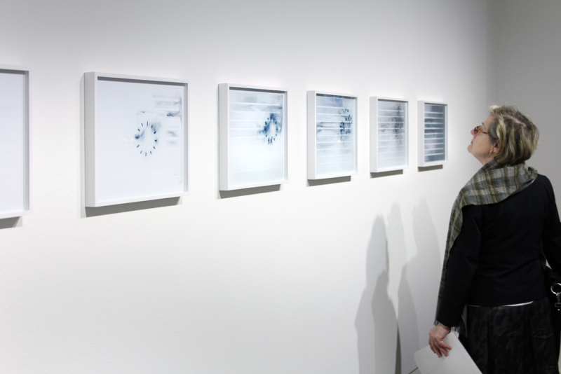  Eine Frau steht vor einer Reihe von weißgerahmten Fotografien, die ein Ziffernblatt aus Oktopustinte in einem schmelzenden Eisblock zeigen.