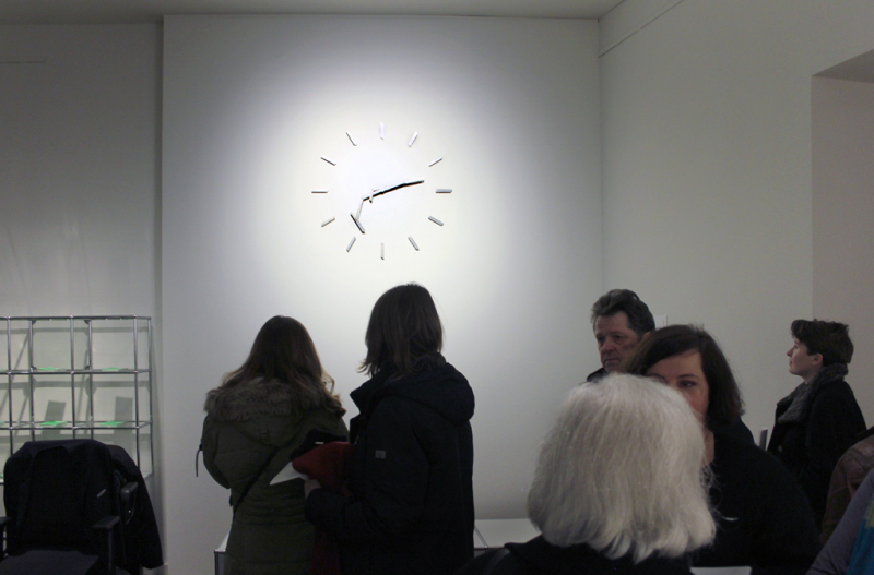  Eine Gruppe von Menschen vor einer hoch an der Wand angebrachten Uhr aus Cuttermesserklingen mit Doppelpendelzeigern.