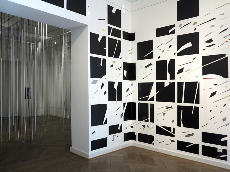  Im HintergrunÜber den Eckbereich des Eingangsraums der Galerie erstreckt sich eine abstrakte Wandzeichnung aus Farbklebeband und Garn. Schwarze Quadrate sind die Grundlage der dynamischen Komposition und reflektieren die Eingangsstruktur der Galerie.