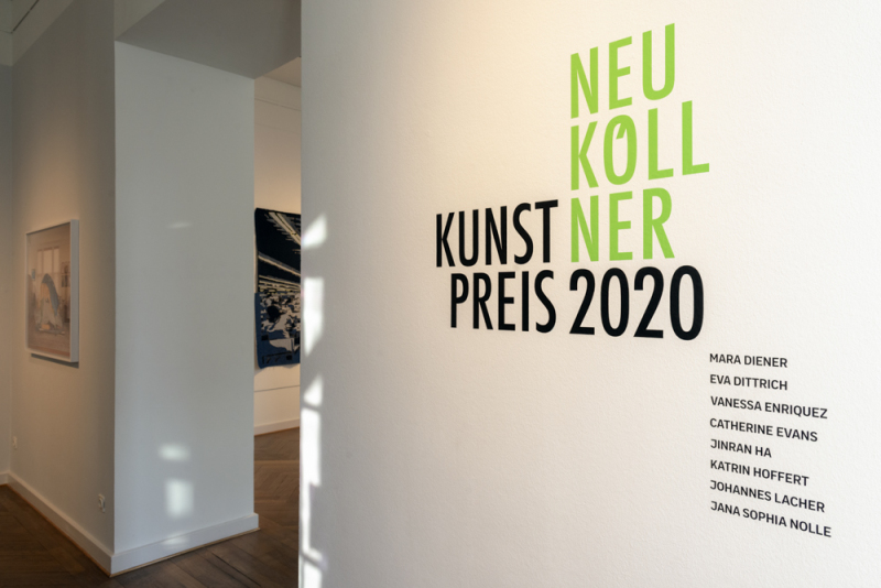 Das Logo zum Neuköllner Kunstpreis 2020 ist in grüner und schwarzer Schrift auf der Wandfläche in der Ausstellung
