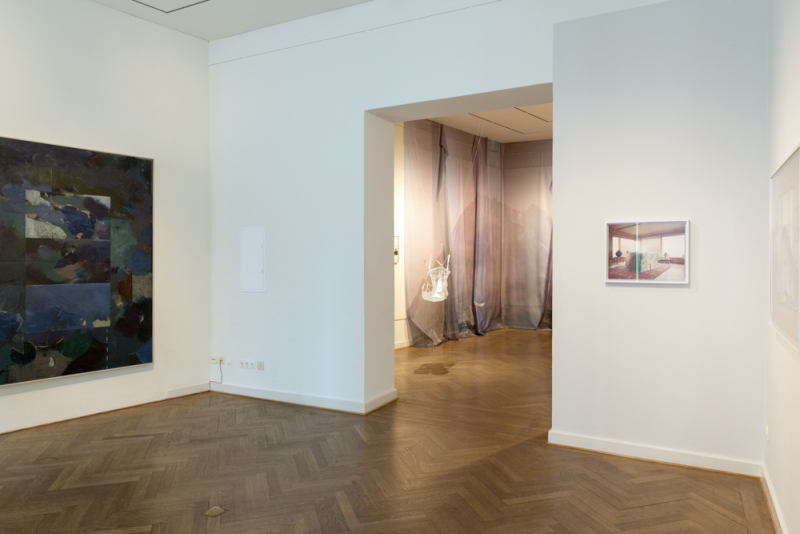 Links hängt eine dunkelblaue Malerei, mittig Gurtzeug und Fallschirmseide und rechts eine Fotografie von einem Wohnzimmer
