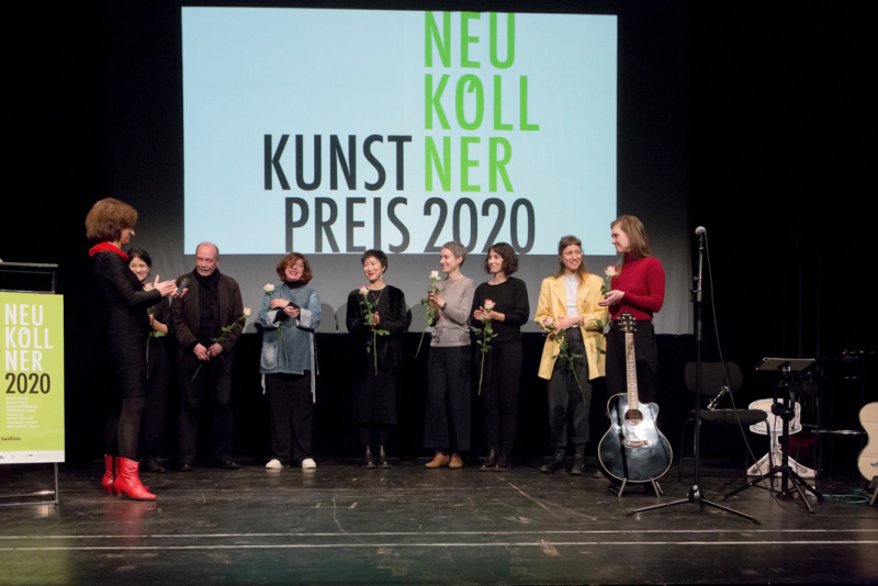Die 8 Nominierten Künstler*innen stehen auf der Bühne vor der Projektion der Neuköllner Kunstpreis 2020 Schrift mit jeweils einer Rose in der Hand