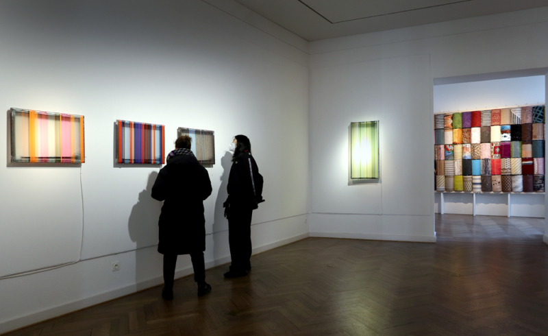 Zwei Besucher*innen stehen vor drei kleinformatigen Werken "Glatt, Glatt, Glatt", die an der Wand hängen