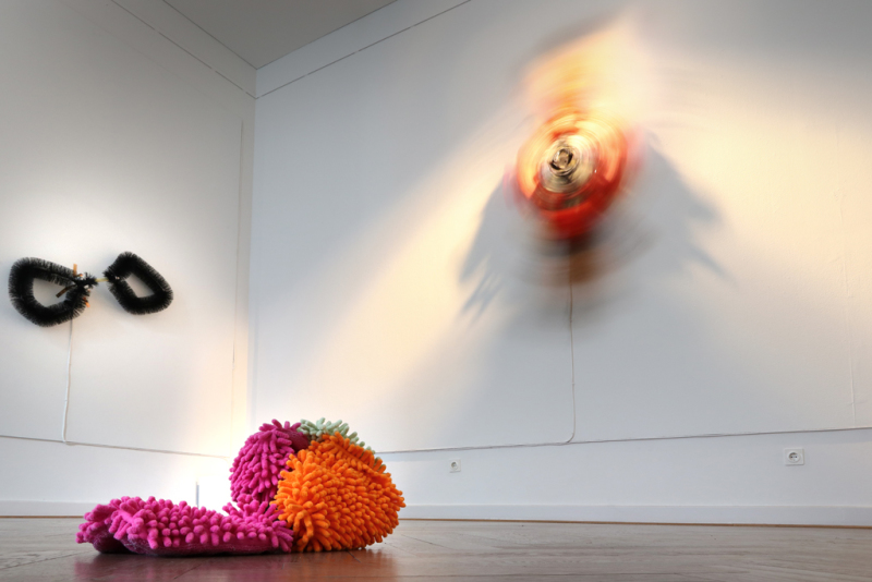 Im Vordergrund ein kleines Objekt der Serie Soft Power: Play time (I – II), bestehend aus einem Spielzeugauto und Putzutensilien und an der Wand ein sich bewegendes Objekt