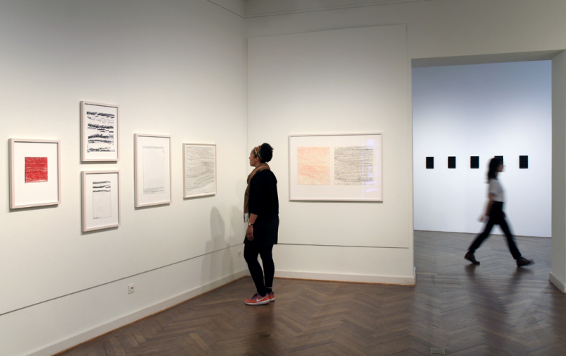 eine Besucherin steht vor den Zeichnungen von Harald Stoffers und eine Besucherin läuft in dem Raum mit den Werken von Etienne Dietzel