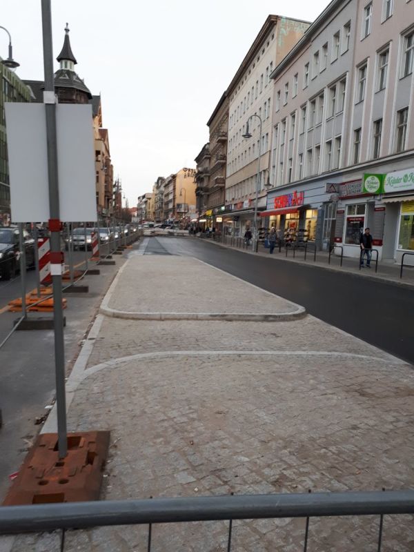 Foto: frisch geteerte Straße und neu angelegte Verkehrsinsel durch Bauzaun von der Straße mit Geschäften getrennt