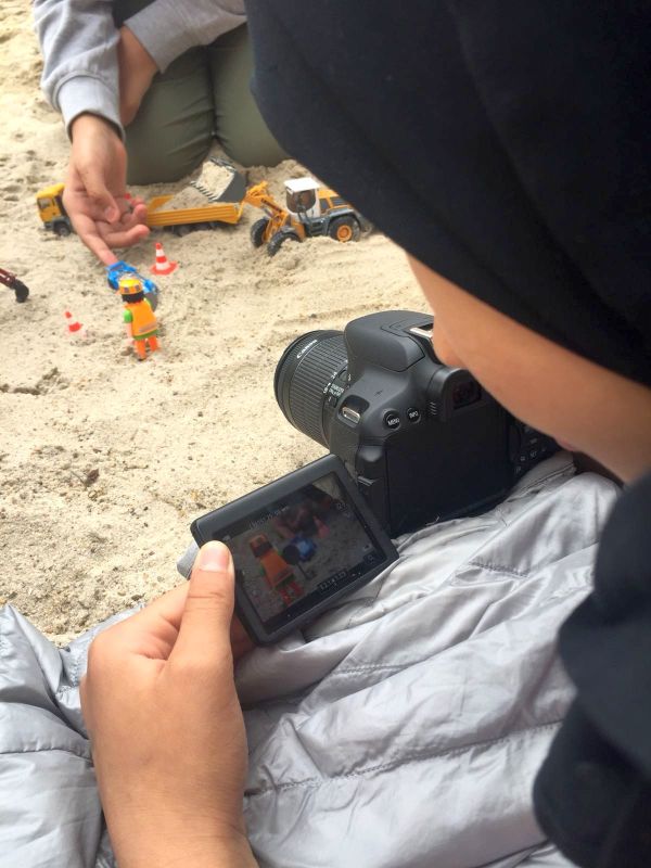 Blick über die Schulter eines Mädchens, das im Sand eine Spielzeugbaustelle filmt