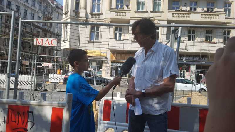 Foto eines Jungen mit einem Mikrofon in der Hand, der einen Mann interviewt, im Hintergrund ein Bauzaun und Häuser