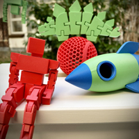 3D-Druckobjekte: links ein roter Roboterm in der Mitte ein roter Ball, rechts eine blau-grüne Rakete 