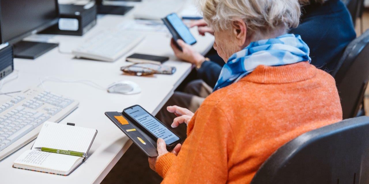 Kursszene - eine ältere Frau mit dem Smartphone in der Hand, sitzend im Computerraum