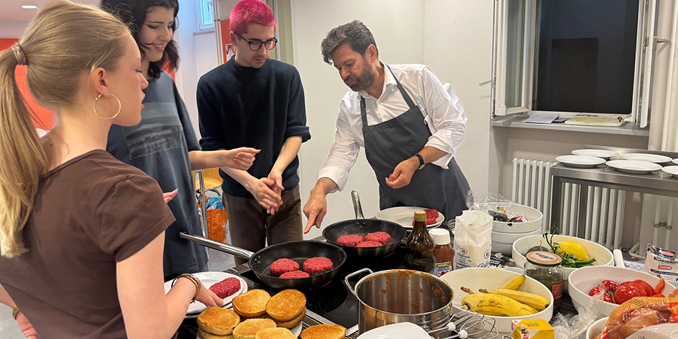Kochkurs-Szene mit Jugendlichen und Kursleiter Michael Adrians: Das Braten von Burgerfleisch wird erklärt