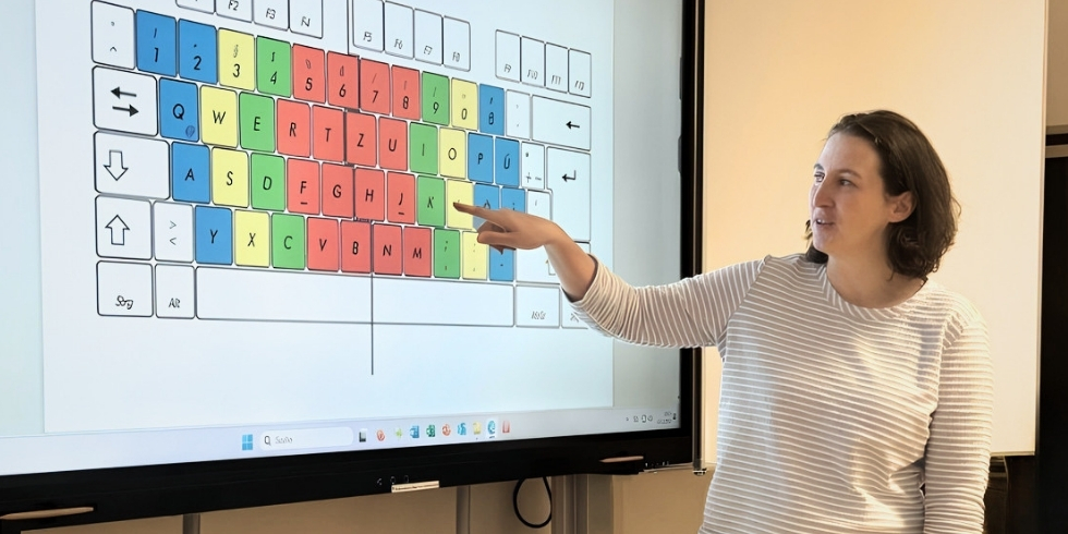 Kursleiterin Neig steht vor dem Whiteboard. Sie zeigt auf eine abgebildete Tastatur. Die Tasten sind unterschiedlich farbig.
