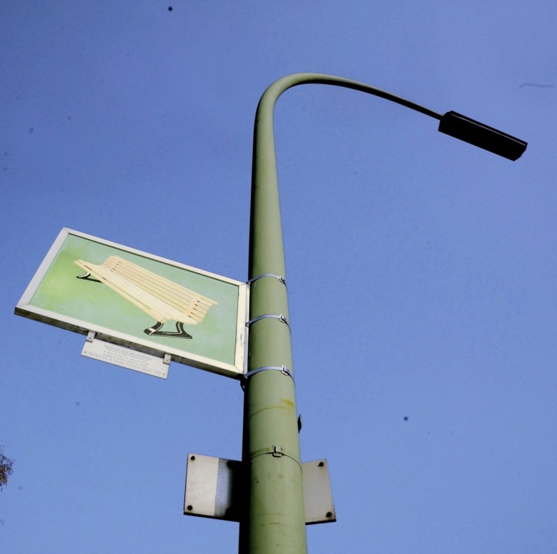 An dieser Laterne ist auf der einen Seite des Schildes eine gelbe Parkbank zu sehen
