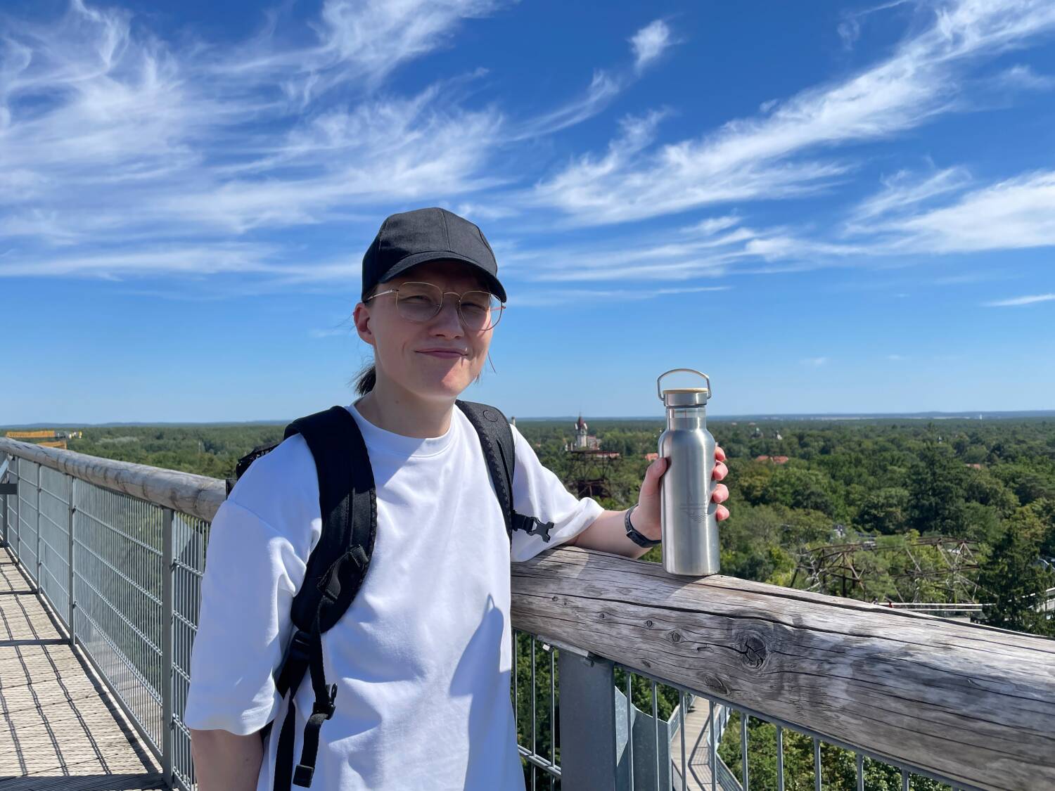 Person auf einem Baumwipfelpfad mit Edelstahlflasche in der Hand und Basecap auf dem Kopf