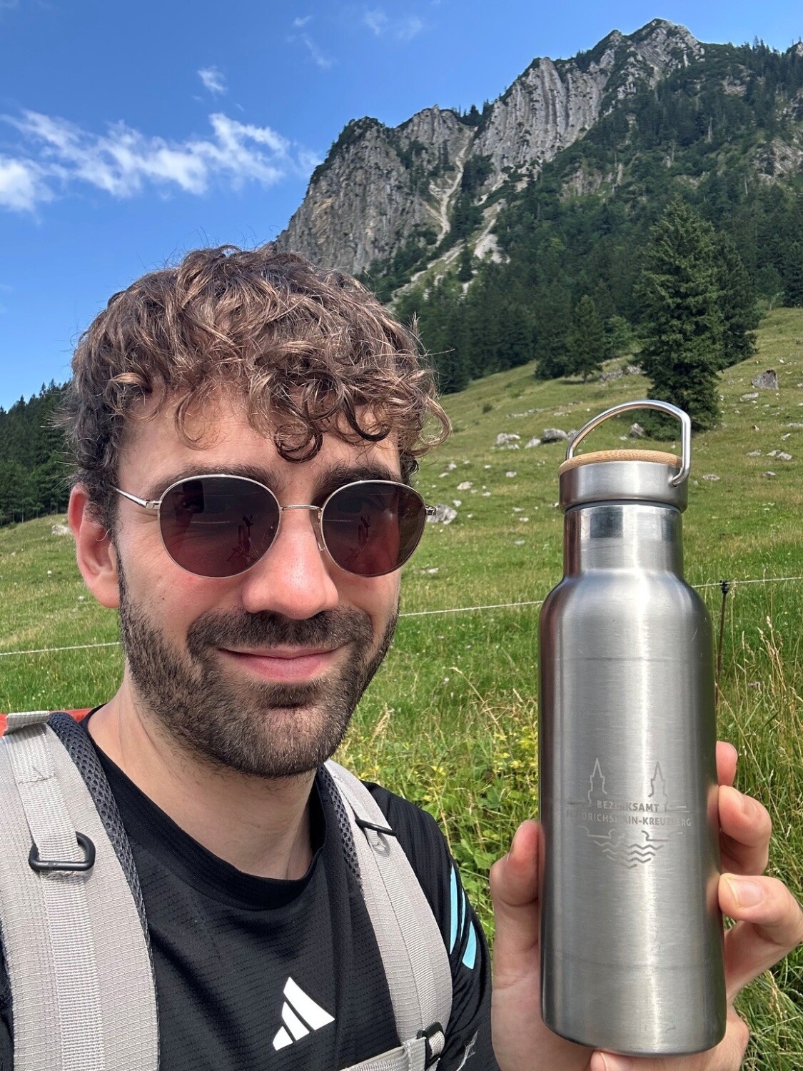Lächelnde Person mit Sonnenbrille hält Edelstahlflasche mit Gravur in Natur hoch.