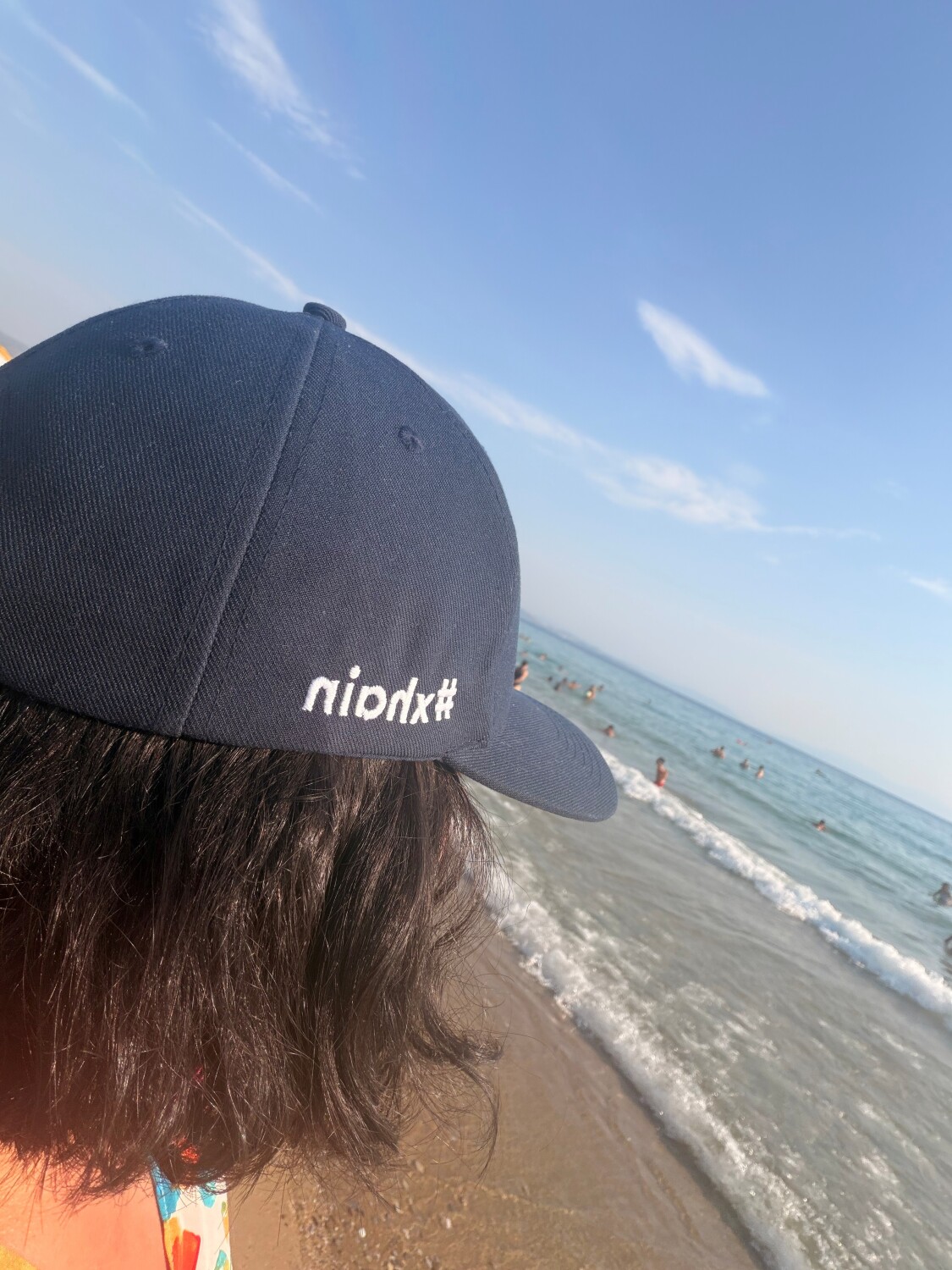 Person mit dunklem Haar und schwarzer Kappe mit weißer Schrift am Strand mit Blick auf das Meer und den Himmel