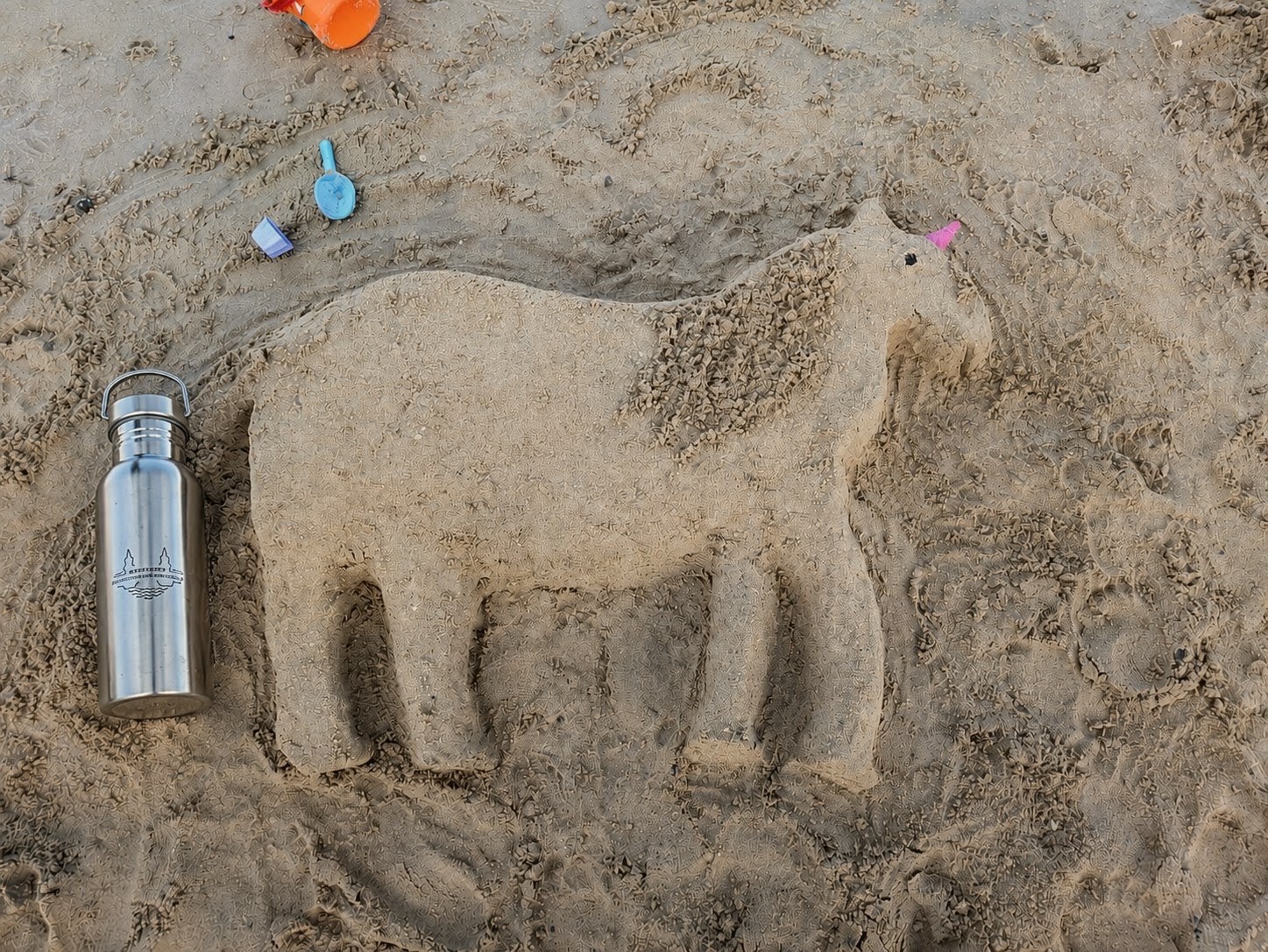 Sandfigur eines Elefanten mit einem rosa Strohhalm im Rüssel neben einer silbernen Trinkflasche und buntem Sandspielzeug auf Sandboden