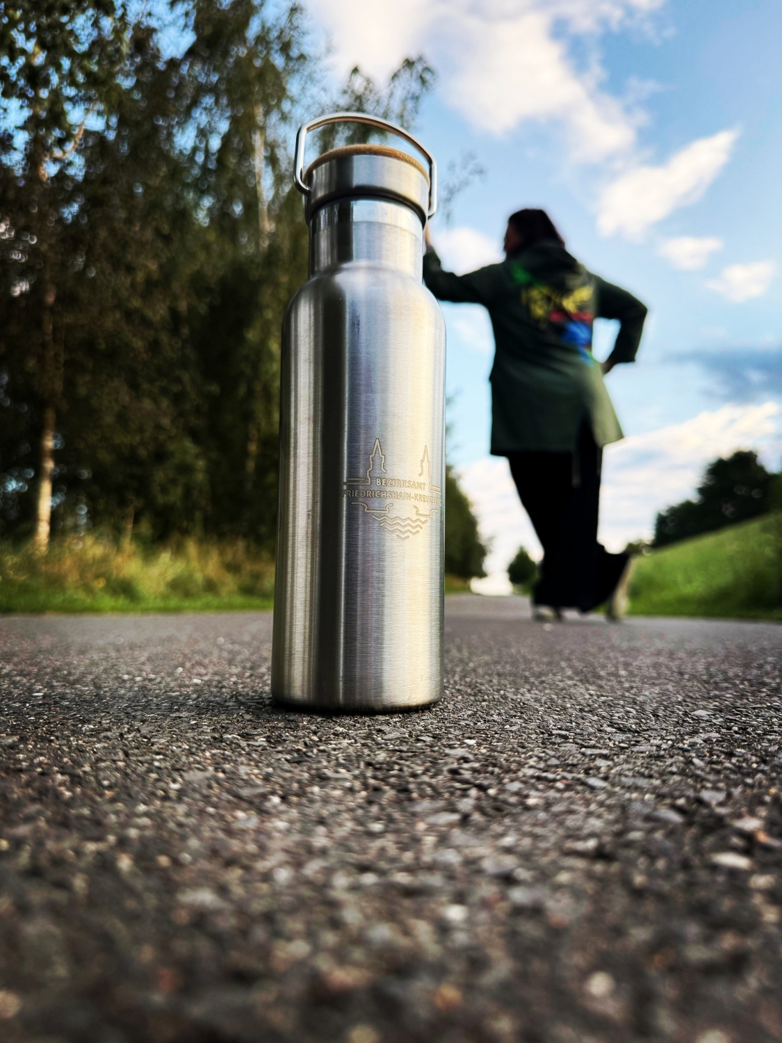 Edelstahlflasche mit Gravur steht auf Asphaltweg, im Hintergrund unscharfe Person mit grünem Mantel vor bewölktem Himmel und Bäumen