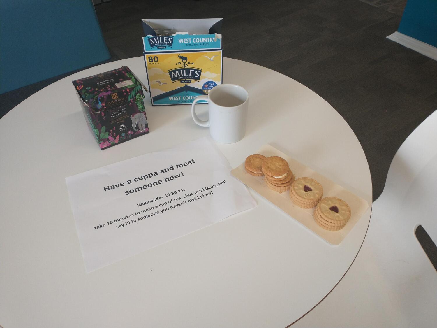 Kekse und eine Kaffeetasse auf einem Tisch, dazu ein Schild mit der Aufschrift "Have a cup and meet someone new"