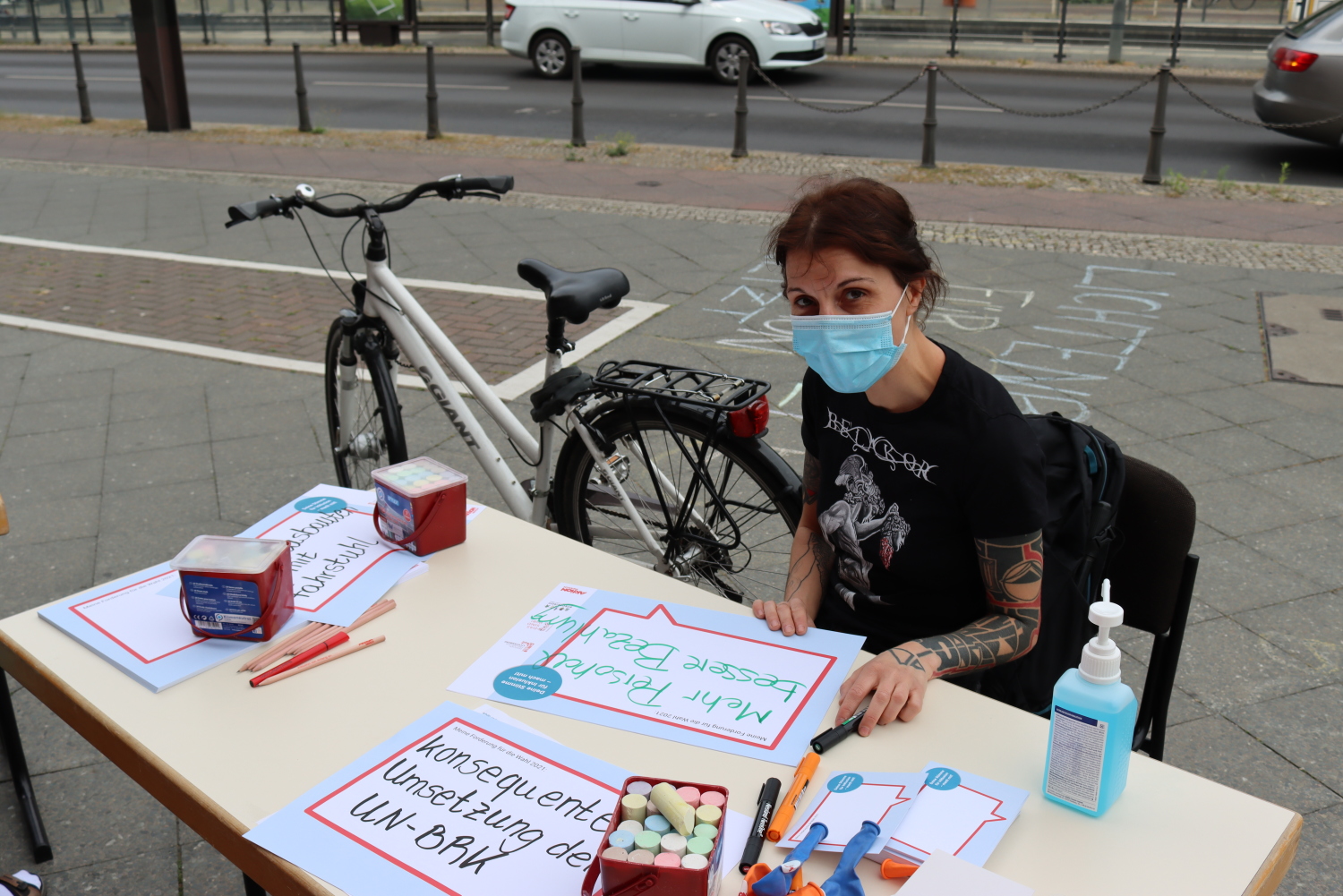 Eine Besucherin sitzt an einem Tisch neben dem Rathaus. Auf dem Tisch liegen Stifte, Kreide, Postkarten, Plakate und Desinfektionsmittel. Auf dem Plakat der Besucherin steht: "Mehr Personal, bessere Bezahlung".