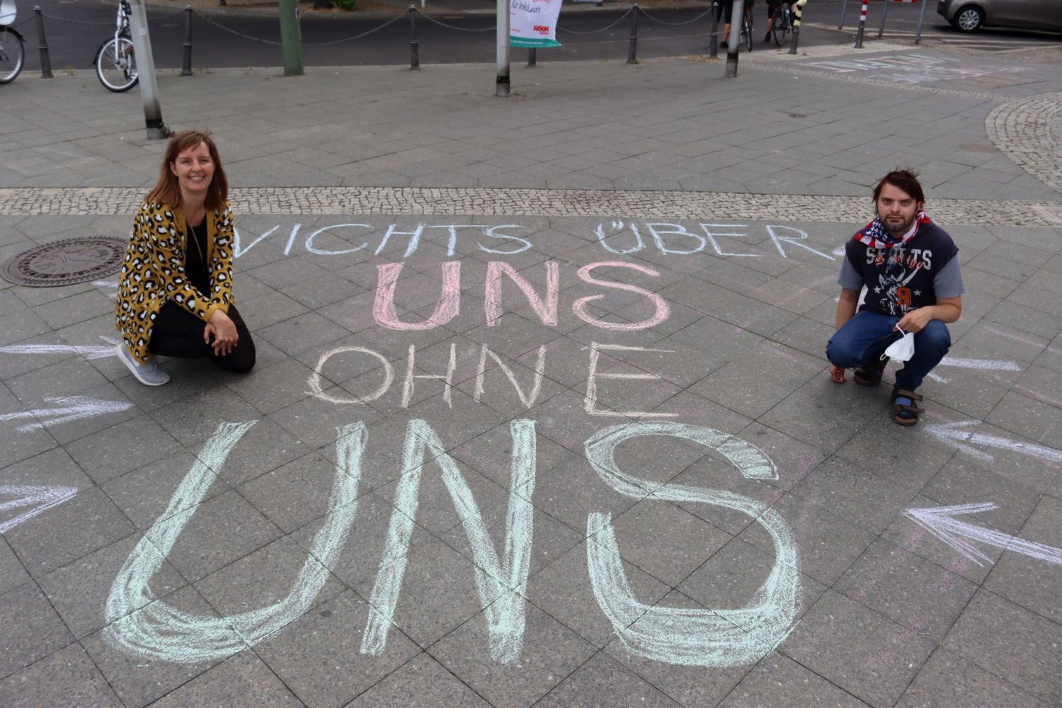 Katja Tieks (links) und Florian Leue (rechts) hocken neben dem mit Straßenkreide gemalten Spruch: "Nichts über uns ohne uns"