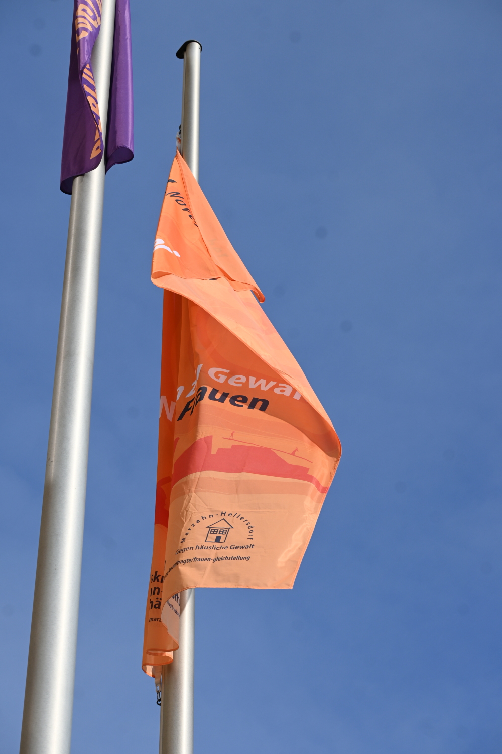 Zwei Fahnen hängen am Fahnenmast. Eine ist lila, eine ist orange. Auf der organenen ist der Text "Gewaltfrei leben" zu erkennen.