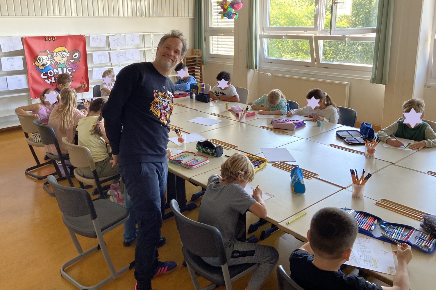 Workshopleiter Steffen Hand steht vor dem Gruppenarbeitstisch, an dem mehrere Kinder zeichnen.