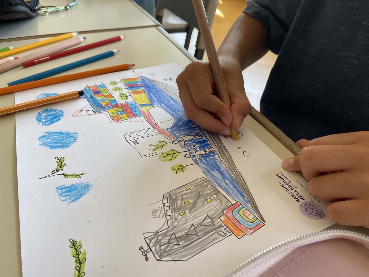 Nahaufnahme einer Kinderhand, einen Stift haltend, über dem eigenen Comicentwurf. Zu sehen ist ein bunter Platz mit Menschen, Bäumen und einer Eisfläche zum Schlittschuhlaufen.