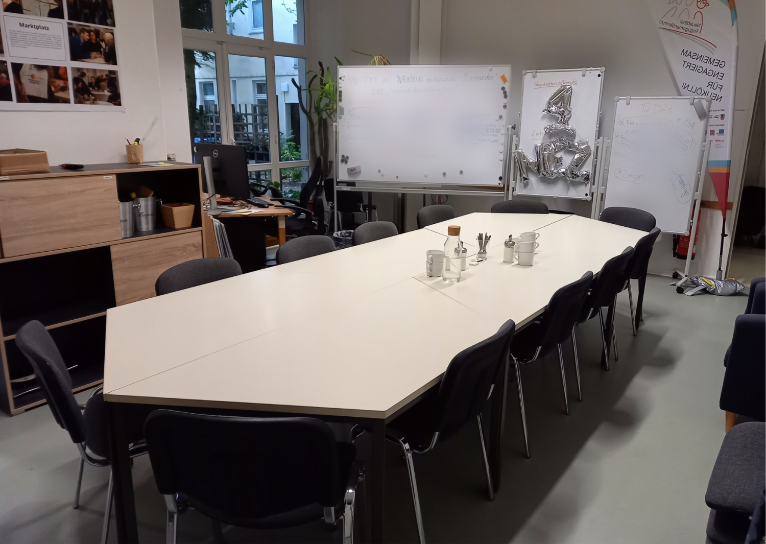 Seminarraum mit eckigem Tisch und Stühlen und Whiteboards, Flipcharts und Regalen im Hintergrund.