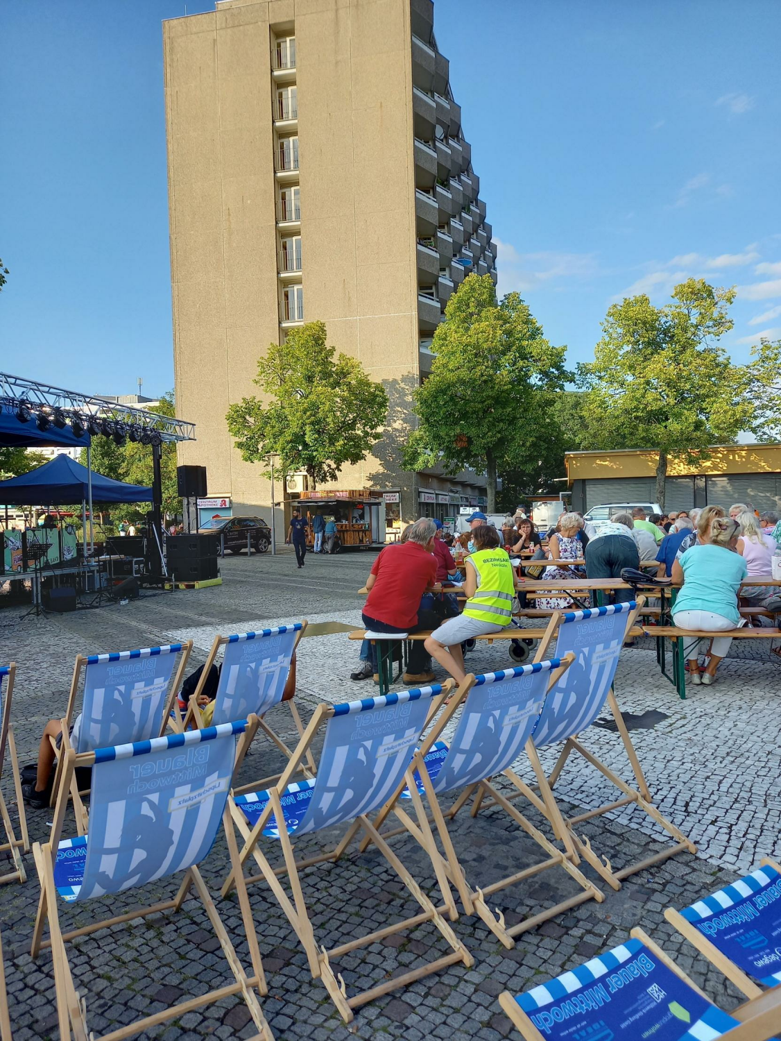 Stühle mit dem Logo des Blauen Mittwoch im Vordergrund. Im Hintergrund unterhält sich eine Person mit gelber Warnweste mit Menschen, die auf Bierbänken sitzen.