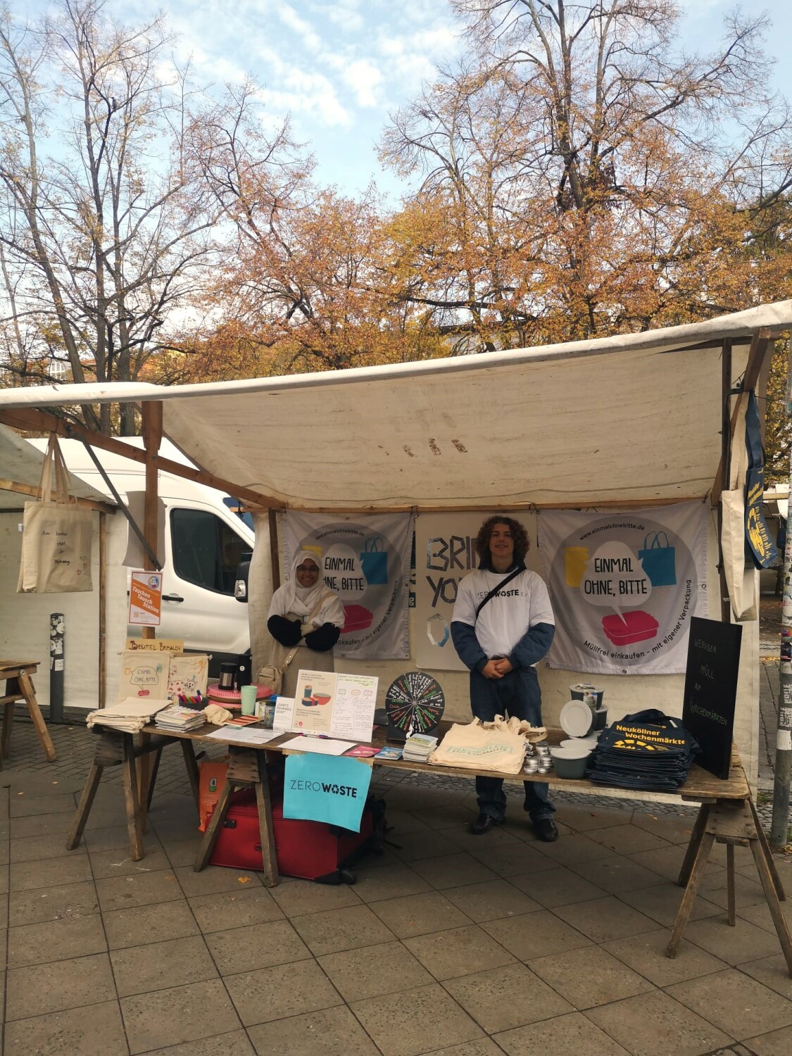 Ein Wochenmarktstand mit Informationsmaterial zum Thema Zero Waste. Auf dem Tische des Standes liegen Beutel, Scheibblöcke, Taschenaschenbecher und andere Merch Gegenstände. Zwei Personen stehen hinter dem Tisch am Stand. Im Hintergrund sind Bäume und blauer Himmel zu sehen.