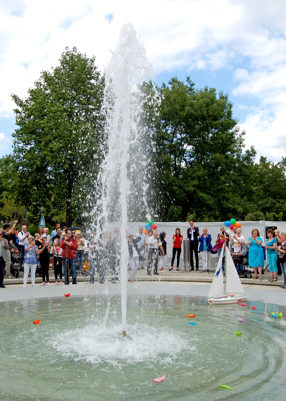 Ein Brunnen mit hoher Wasserfontäne, im Wasser des Brunnens sind kleine bunte Papierschiffe und ein größeres weißes Modellschiff zu sehen, im Hintergrund stehen viele Menschen sie klatschen, einige haben bunte Luftballons in der Hand