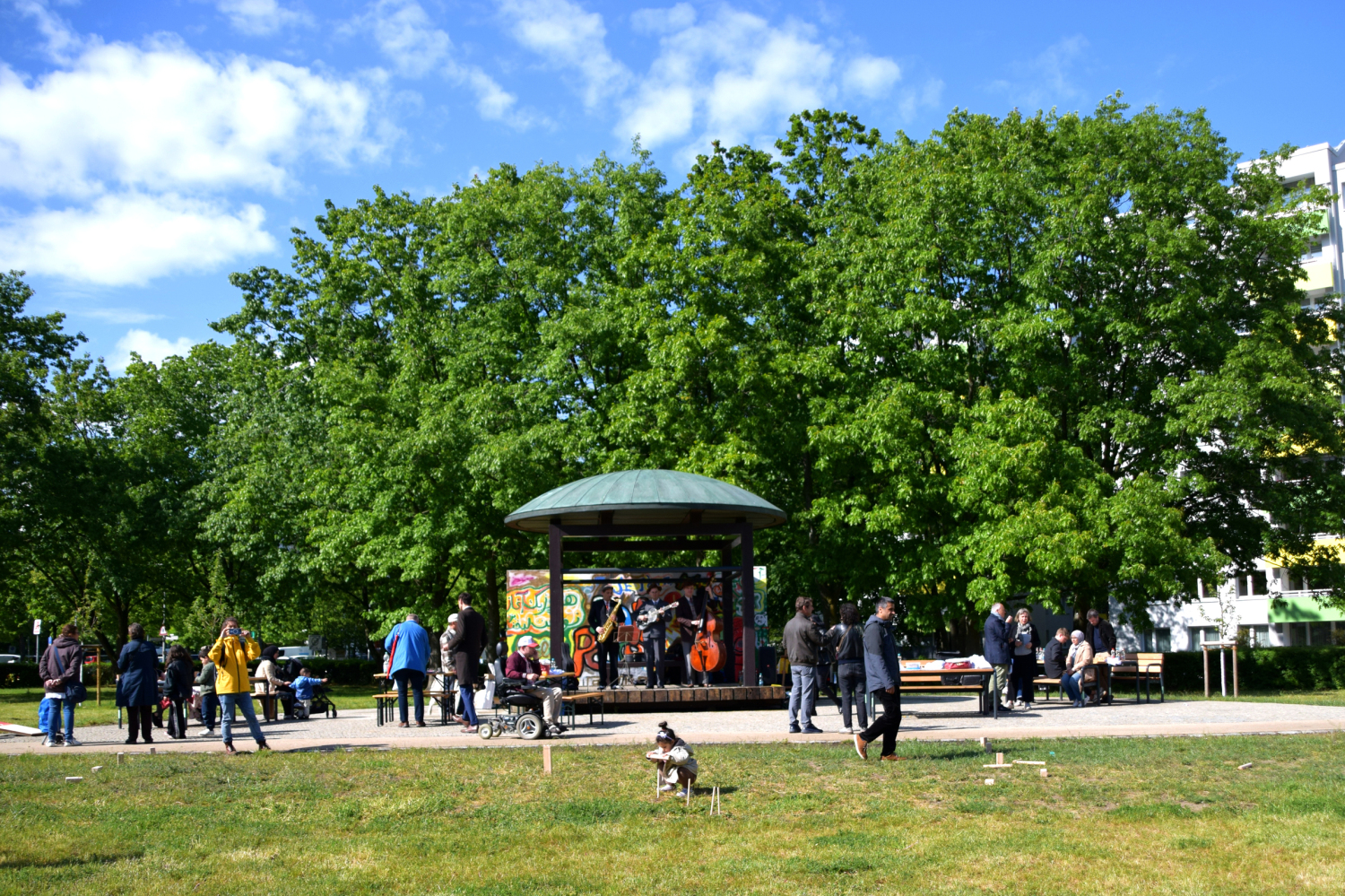 Wiese mit einem festen Pavillion vor einer Wand mit Grafitti. Auf dem Platz vor dem Pavillion stehen Menschen und hören der Band zu die auf der Bühne unter dem Pavillion Musik spielt. Im Hintergrund sind Bäume und blauer Himmel zu sehen.