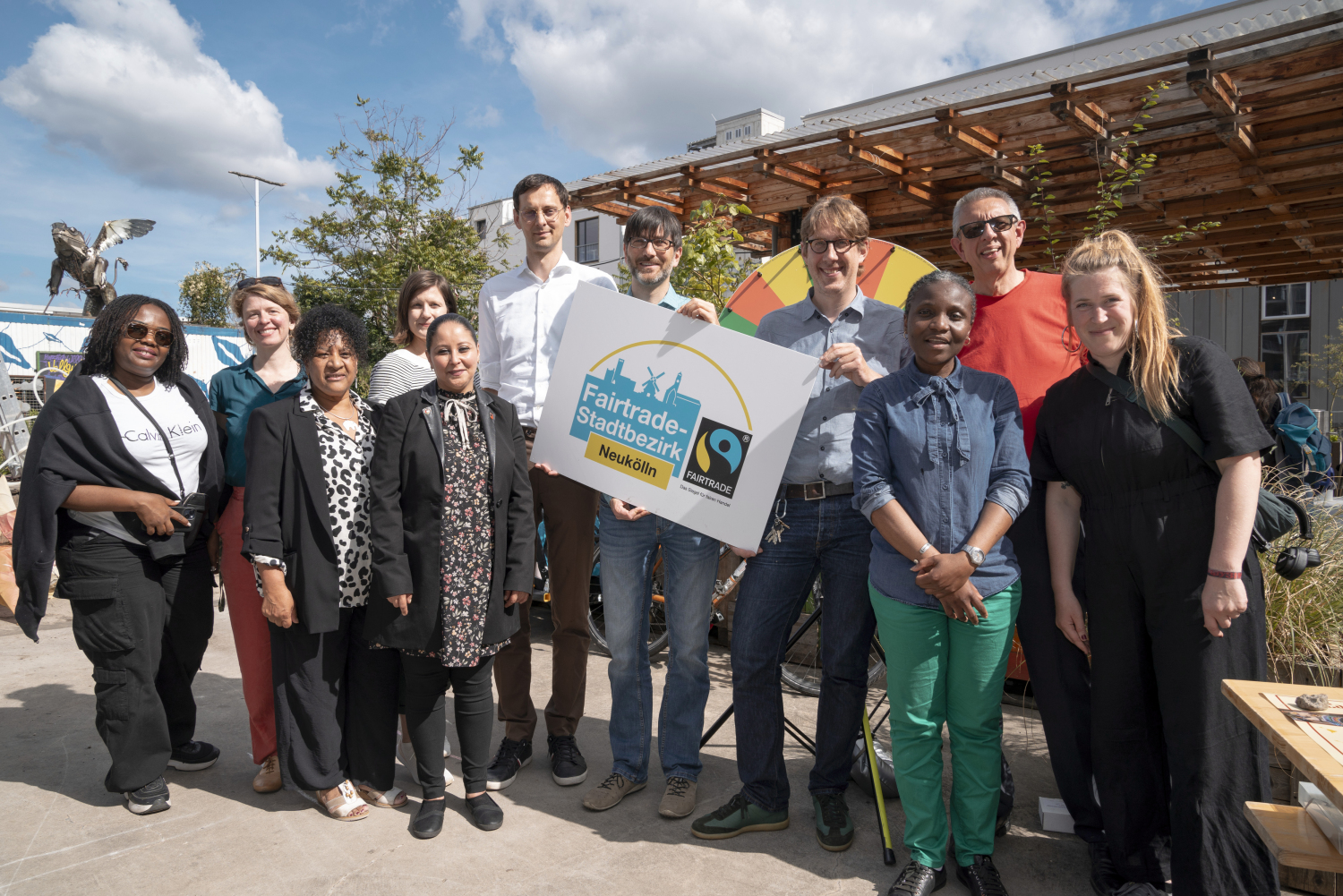 Ein Gruppenfoto mit 11 Personen, darunter der Bezirksbürgermeister Martin Hikel, der Stadtrat Jochen Biedermann und die Nachhaltigkeitsbeauftragte Eva Hein. Eine Person in der Mitte hält ein Schild in der Hand mit der Aufschrift Fairtrade-Stadtbezirk Neukölln und dem Fairtrade Logo. Im Hintergrund ist ein blauer Himmel zu sehen.