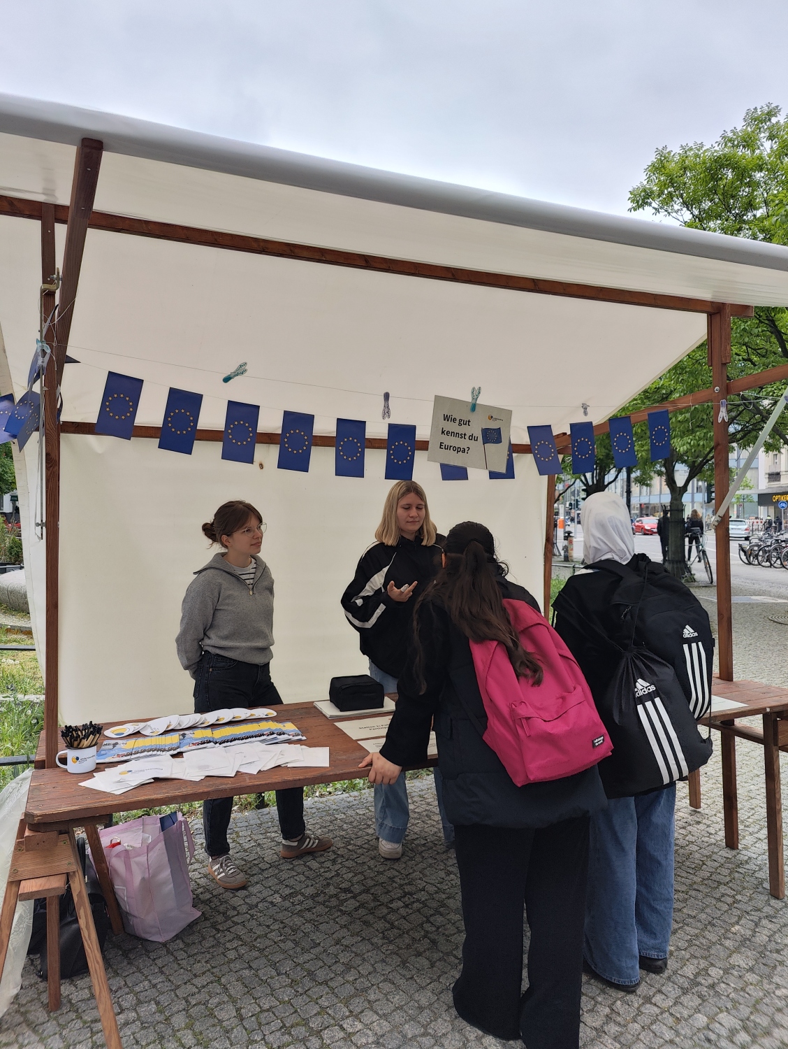 Besuchende unterhalten sich am Info-Stand. Auf dem Stand liegt viel Material. Er ist einer Girlande aus kleinen Flaggen der Europäischen Union gecshmückt .