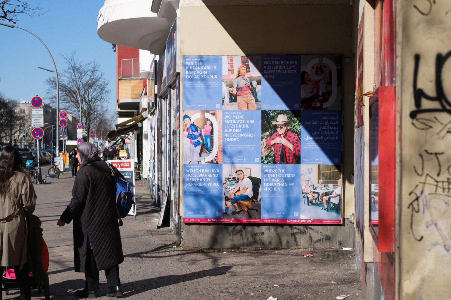 Eine Hauswand mit 6 Verschiednen Plakaten der Kampange Null Müll Neukölln. Die Sonne scheint auf die Plakatwand, links daneben stehen zwei Bürgerinnen. Auf der linken Seite ist ein Bürgersteig zu sehen.