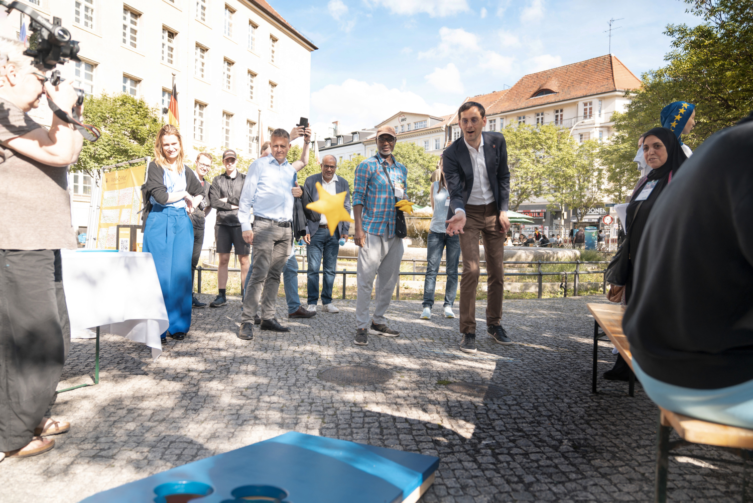 Bezirksbürgermeister Martin Hikel spielt ein Wurfspiel, bei dem Säckchen auf einer Scheibe die am Boden liegt in einem Loch landen müssen. Die Scheibe ist Blau, die Säckchen sind kleine gelbe Sterne. Ein Säckchen befindet dich gerade mitten im Wurf. Um ihn herum stehen mehrere Menschen und schauen zu. Das ganze fand auf dem Rathausvorplatz statt, dieser ist auch im Hintergrund zu sehen.