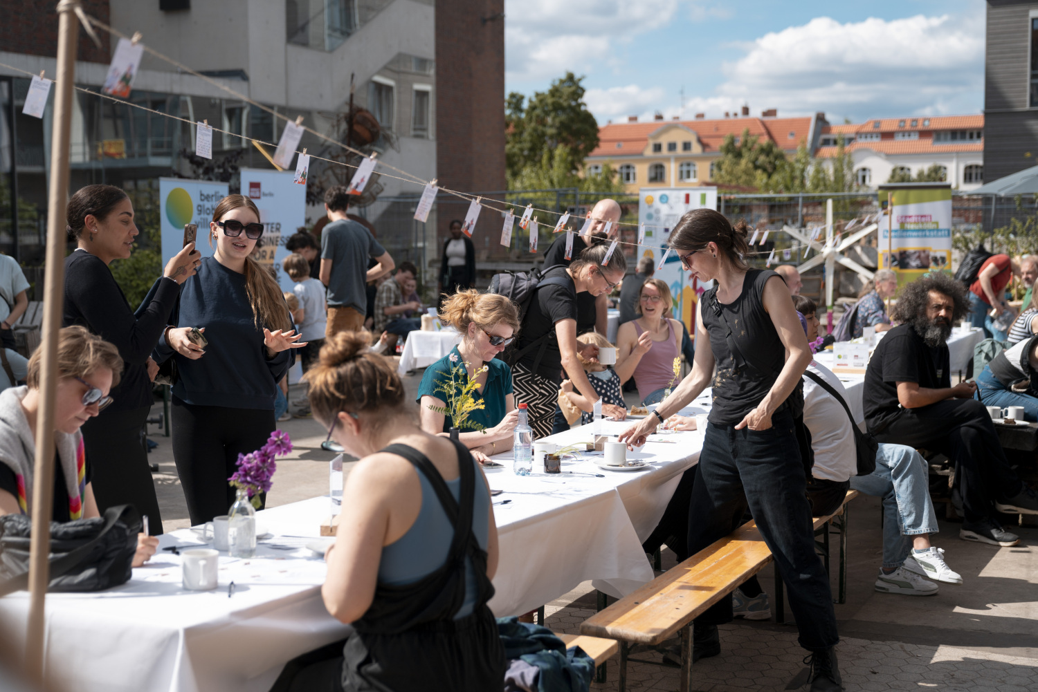 Mehrere Biertischgarnituren stehen zu einer langen Tafel zusammen, sie sind mit weißen Tischdecken bedeckt, umgeben von Menshcen die sich unterhalten und Kaffee trinken. Über den Tischen hängen an einer Schnur kleine Karten. Im Hintergrund sind noch mehr Menschen und Häuser zu sehen.