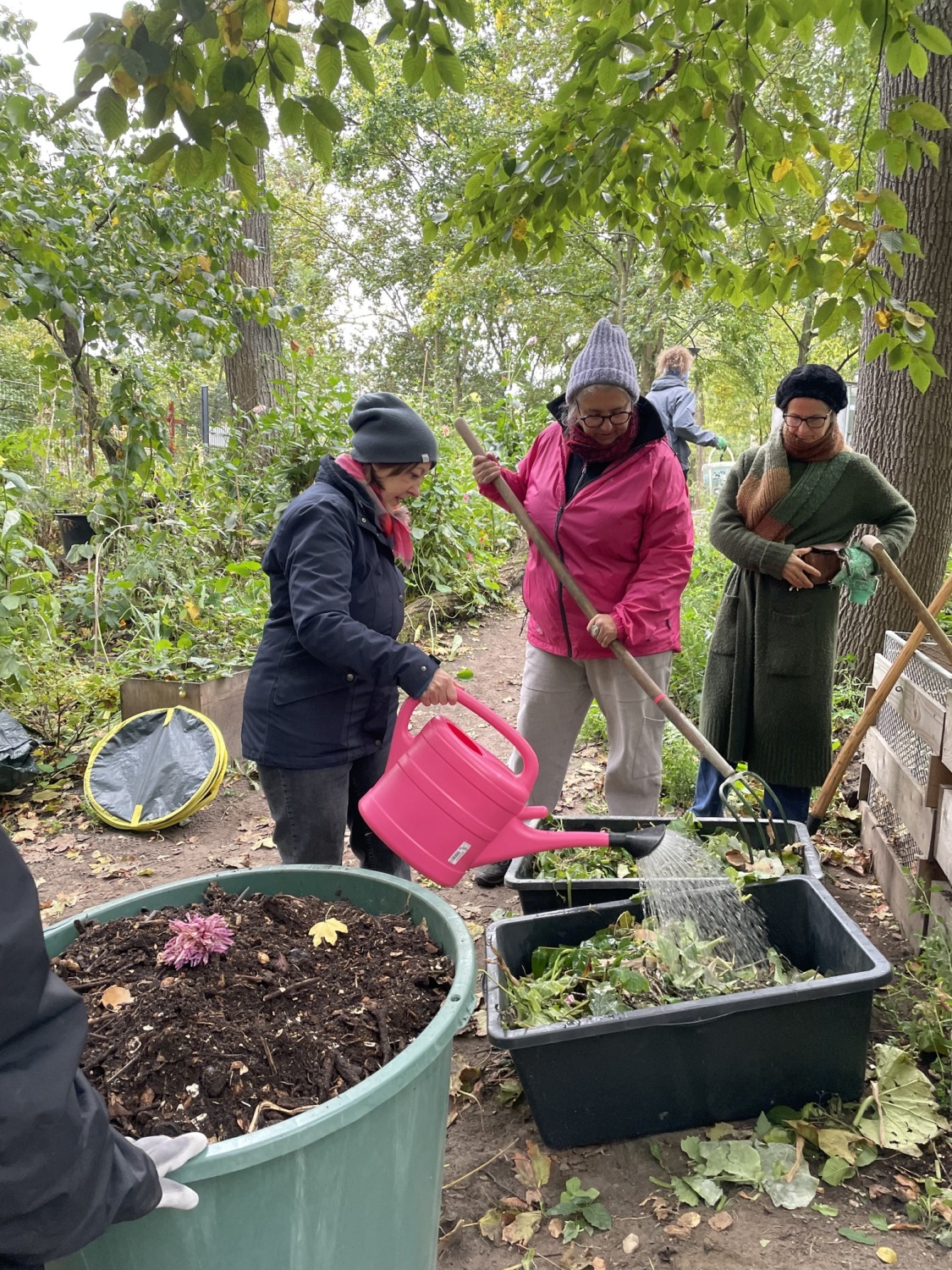 Drei Personen kompostieren Gartenabfälle in einer schwarzen Wanne, eine Person gießt den Gartenschnitt, die zweite Person wendet ihn mit einer Mistgabel, die dritte Person schaut zu