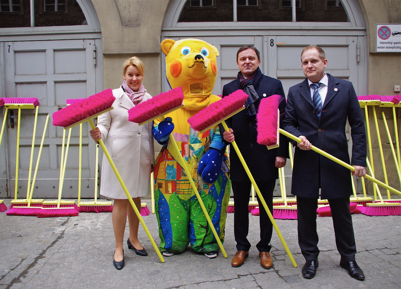 Eine Frau und zwei Männer stehen mit einem Maskottchen des Berliner Bär vor einer Hauswand. Sie halten alle Besen mit gelbem Stiel und pinken Borsten in die Kamera. Im Hintergrund sind ungefähr 20 weitere dieser Besen an eine Hauswand gelehnt.