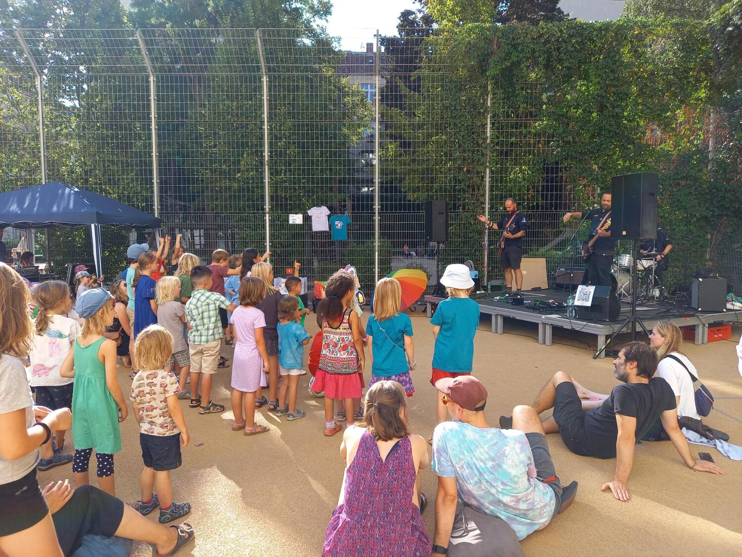Viele Kinder stehen und sitzen auf einem Platz und hören einer Band zu, die auf einer Bühne im Hintergrund Musik spielt.