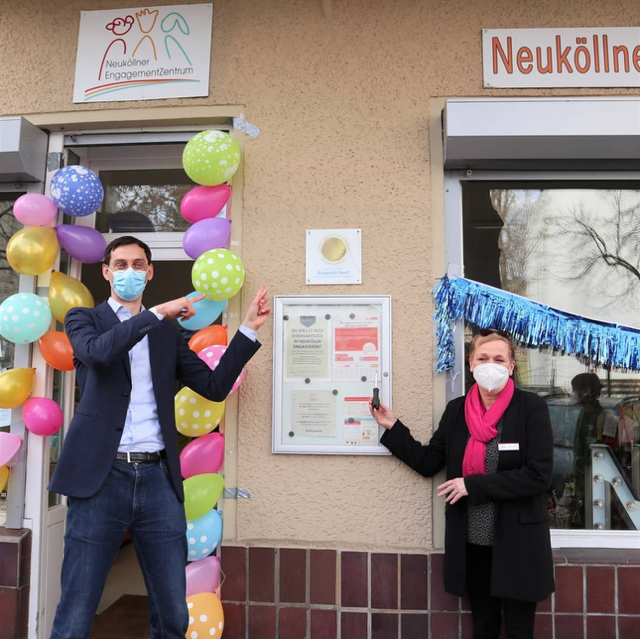 Zwei Personen stehen vor einer Hauswand, zwischen ihnen ist eine Plakette an der Wand angebracht. Sie stehen rechts und links daneben und zeigen auf die Plakette. Die Person auf der linken Seite ist der Bezirksbürgermeister Martin Hikel. Die Hauswand im Hintergrund ist mit Luftballons und einer Girlande geschmückt.