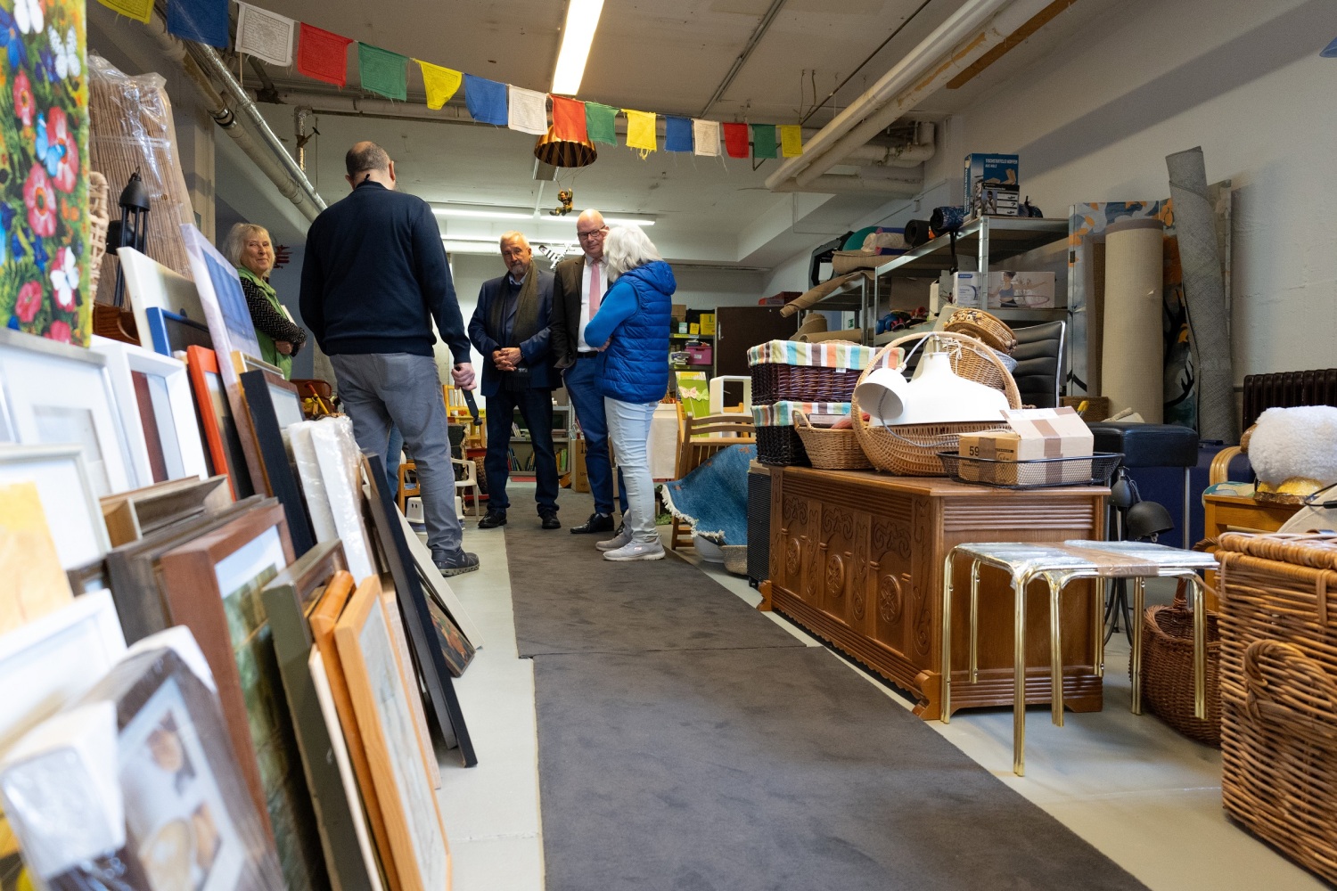 Blick durch einen Gang zwischen Bildern und Möbelstücken zu den Mitarbeitenden des Rumbelbasars im Gespräch mit Herrn Rögner-Francke (mittig) und Herrn Richter (Zweiter von rechts)