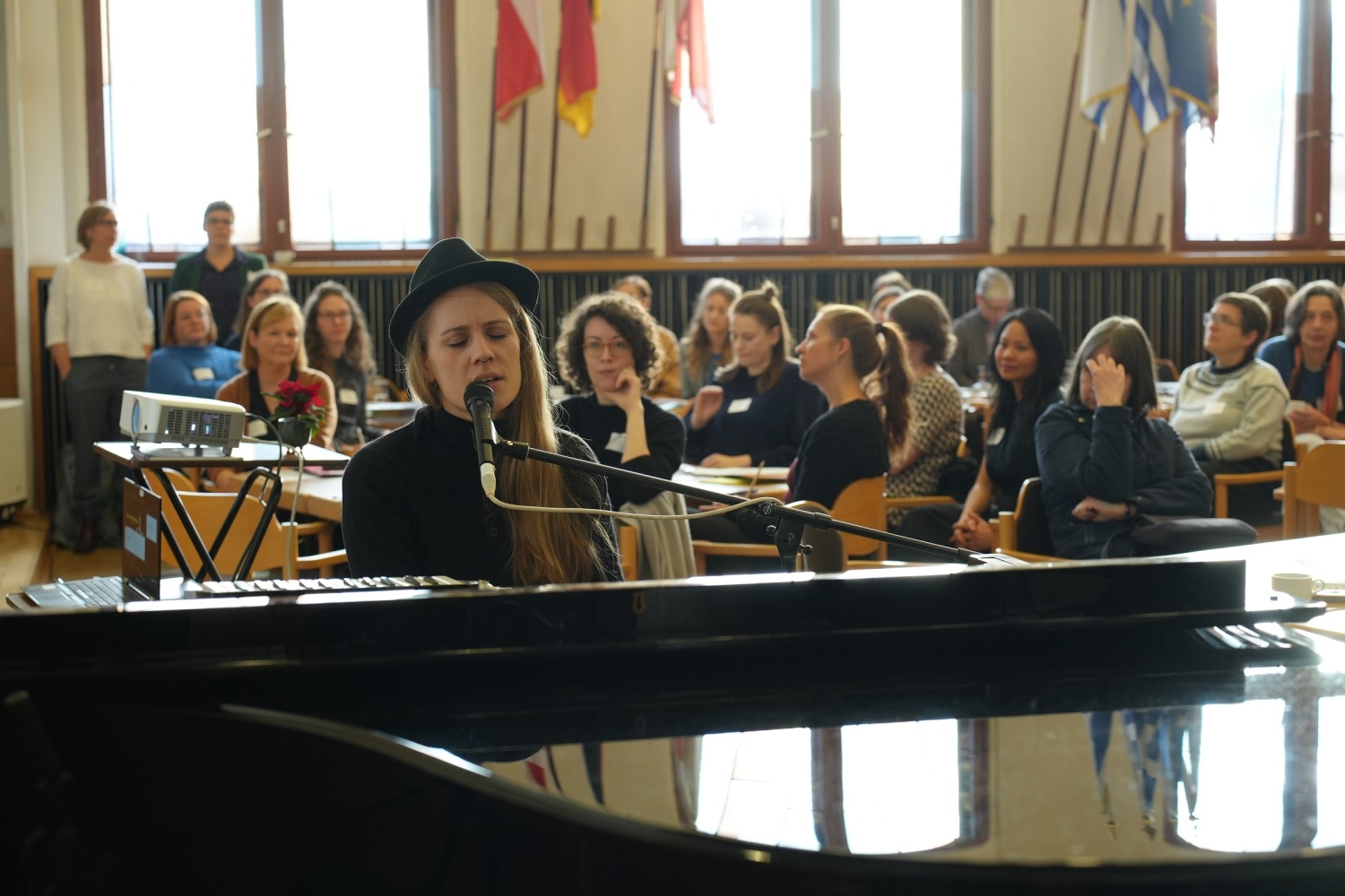 Musikerin Laura Braun singt ihr Lied „Nullsummenspiel“ am Klavier, im Hintergrund hören die Teilnehmenden zu
