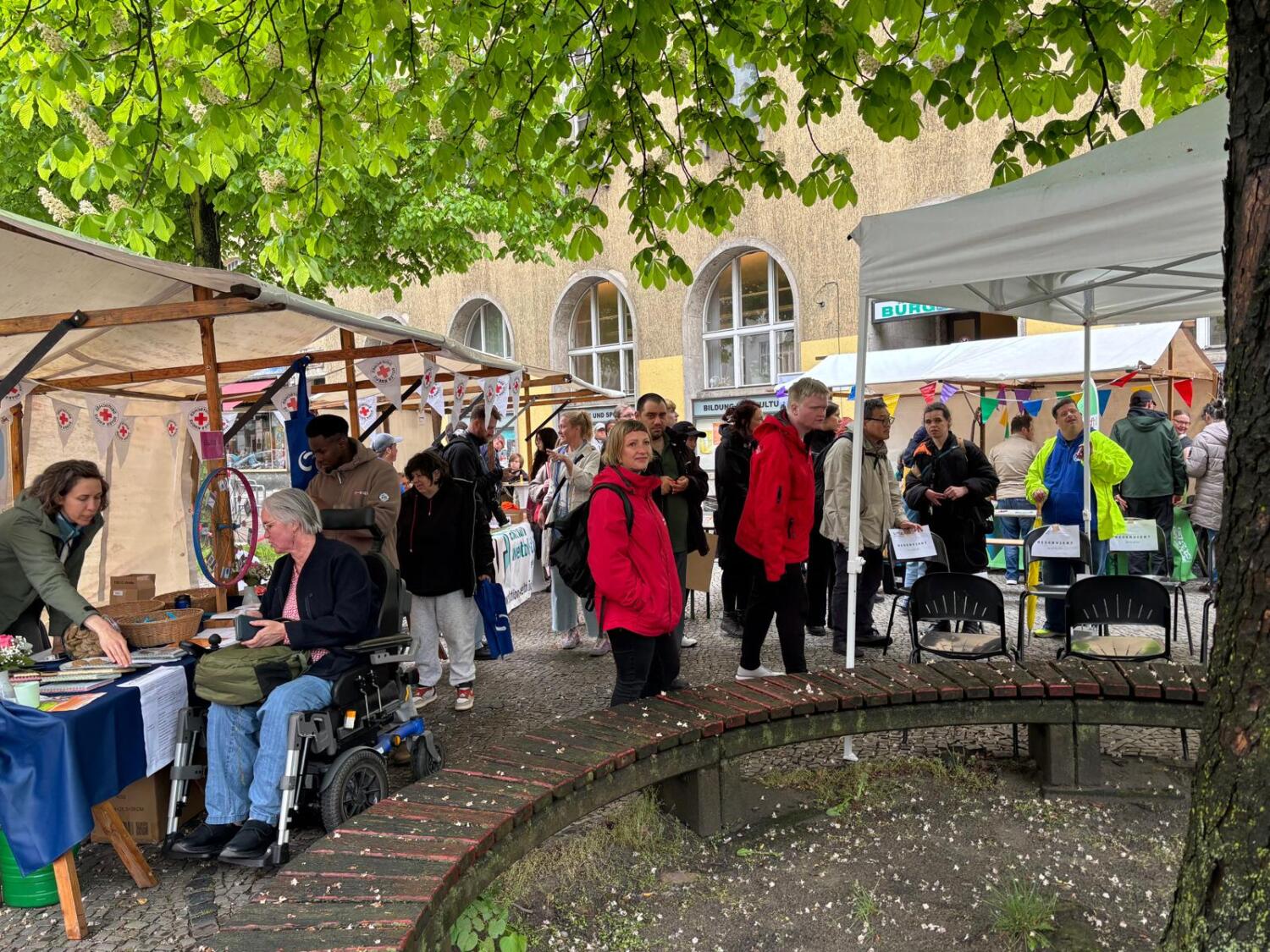 Blick von der Kastanie auf das Fest. Es sind verschiedene Menschen da: Rollifahrerin am Infostand, 2 Menschen in roter Jacke, 1 Person mit Warnjacke. Es scheint verregnet.