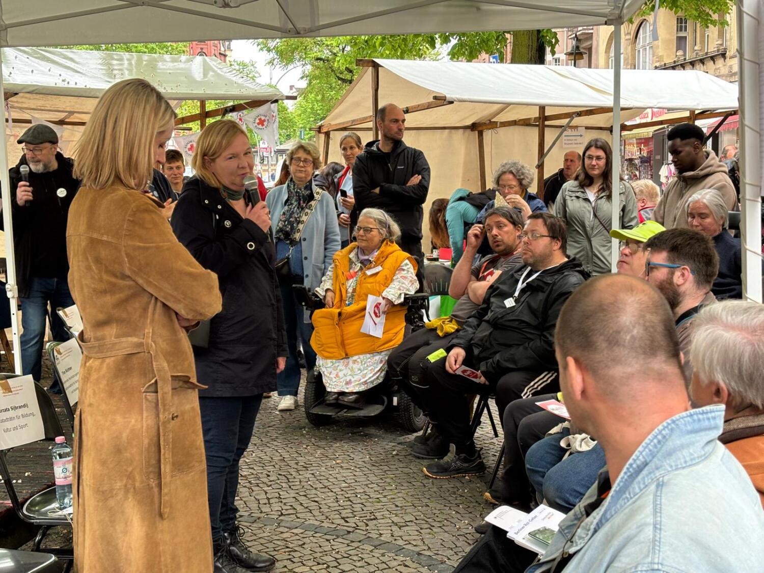 Politik-Talk Mitreden! Inklusion im Bezirk. Im Vordergrund die Stadträtinnen Sijbrandij und Böhm. Im Hintergrund die Beauftragte für Menschen mit Behinderungen Moritz in knallgelber Weste. Im Halbkreis sitzen und stehen viele Interessierte.