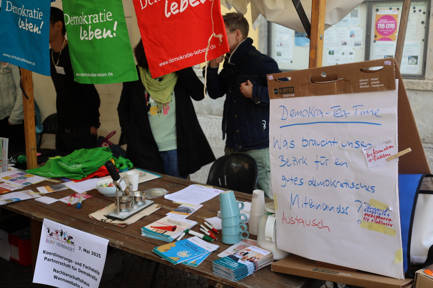 Infostand der Koordinierungs- und Fachstelle Partnerschaft für Demokratie. Es gibt eine Umfrageplakat zum Thema Demokra-TEA-time. Es sind bunte Stoffe (blau, grün, rot) aufgehängt, auf denen steht in weißer Schrift "Demokratie leben!".