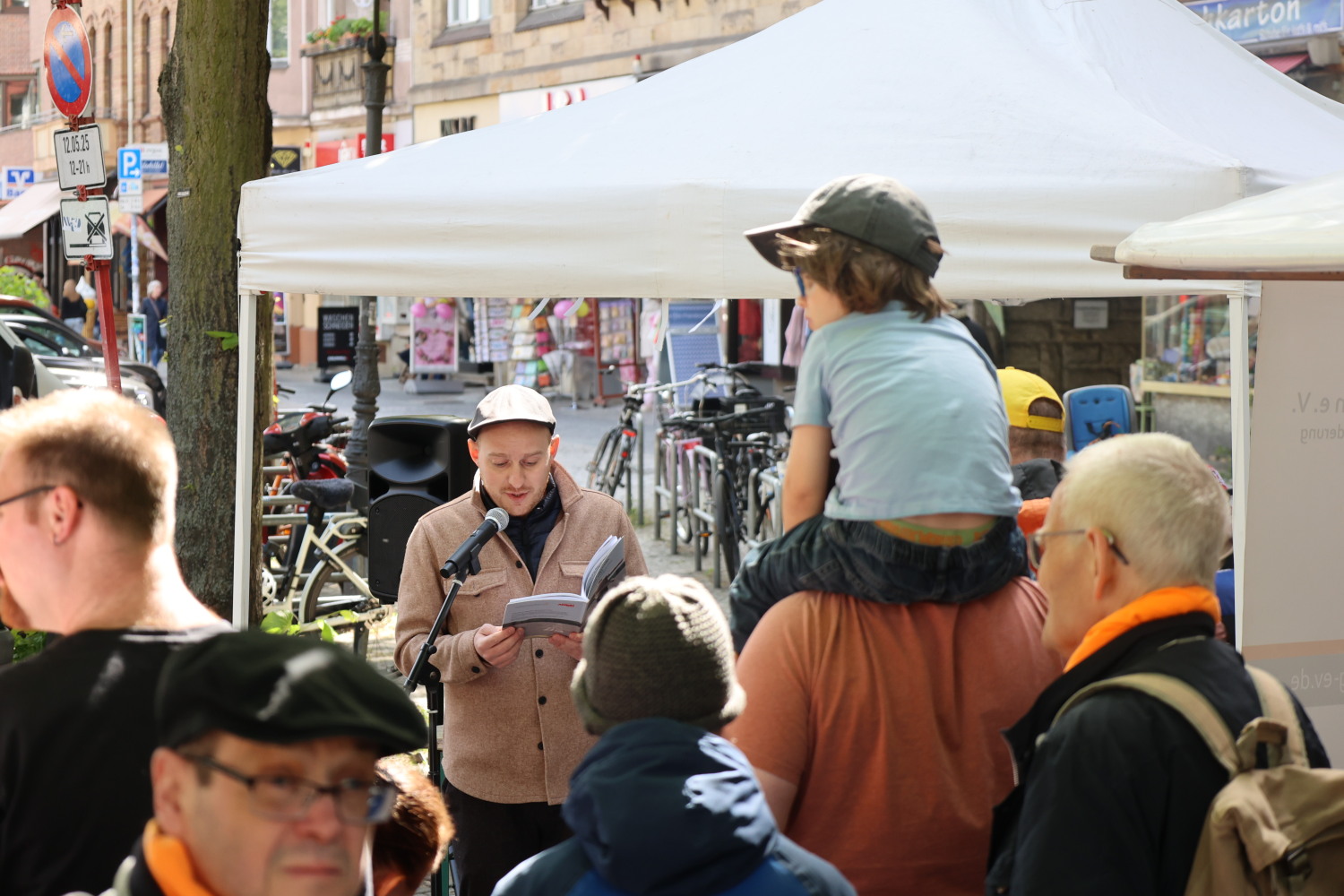 Jemand liest aus dem Buch der Zukunftssicherung Berlin e. V. "Wie ich wurde wer ich bin". Menschen hören zu. Ein Kind sitzt auf den Schultern.